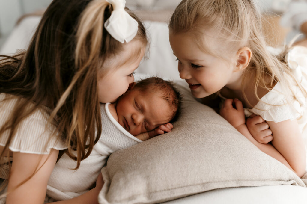 Sisters snuggling newborn on bed in neutral home setting