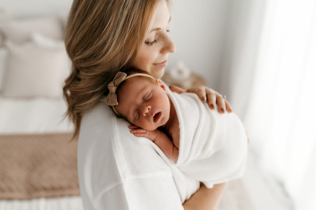 Mom snuggles newborn near the window in natural light.